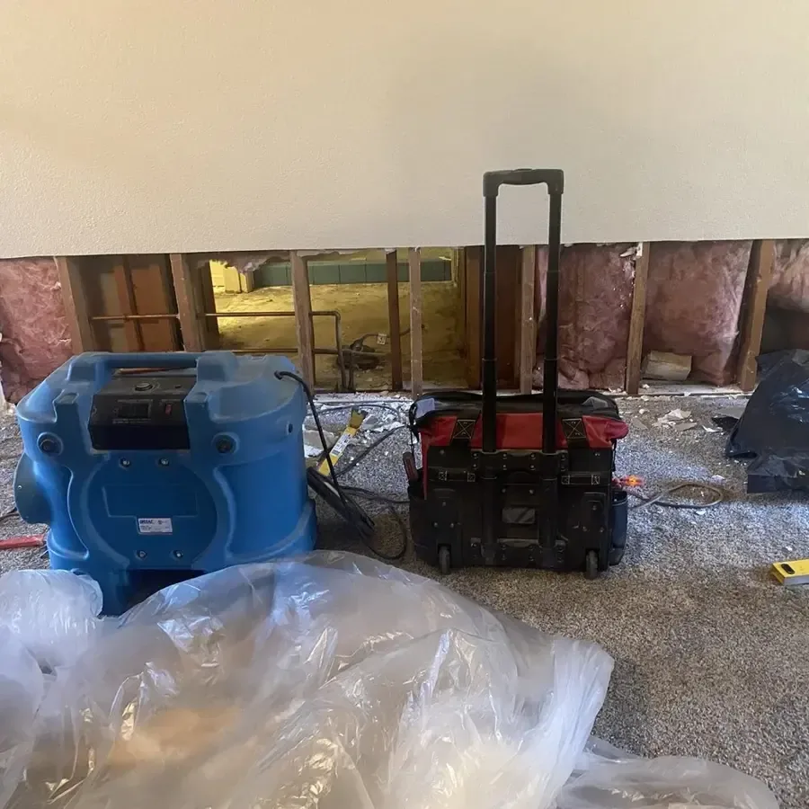 Air movers and a dehumidifier beside a wall opened along the lower framing