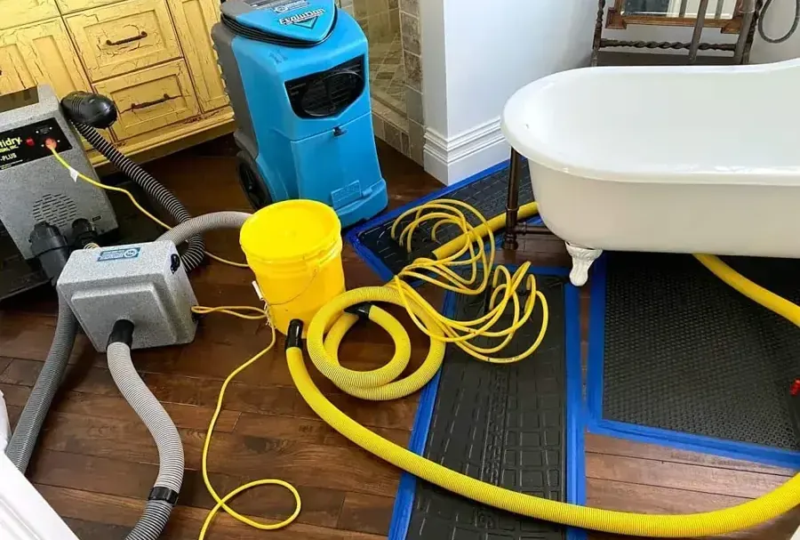 A dehumidifier, floor drying mats and yellow hoses beside a white bathtub